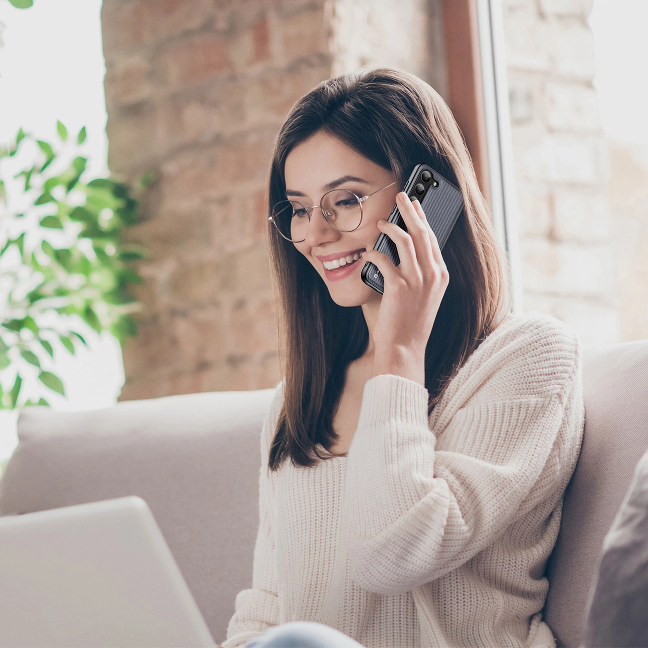 Una donna sorridente con gli occhiali è seduta su un divano, parla al telefono e usa un laptop.