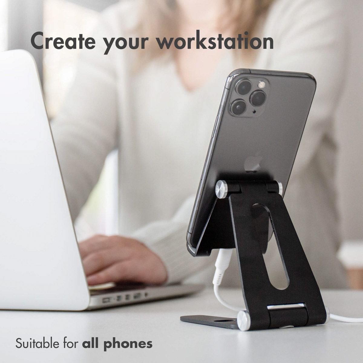 Uno smartphone su un supporto nero accanto a un laptop su un tavolo, con una persona sullo sfondo.