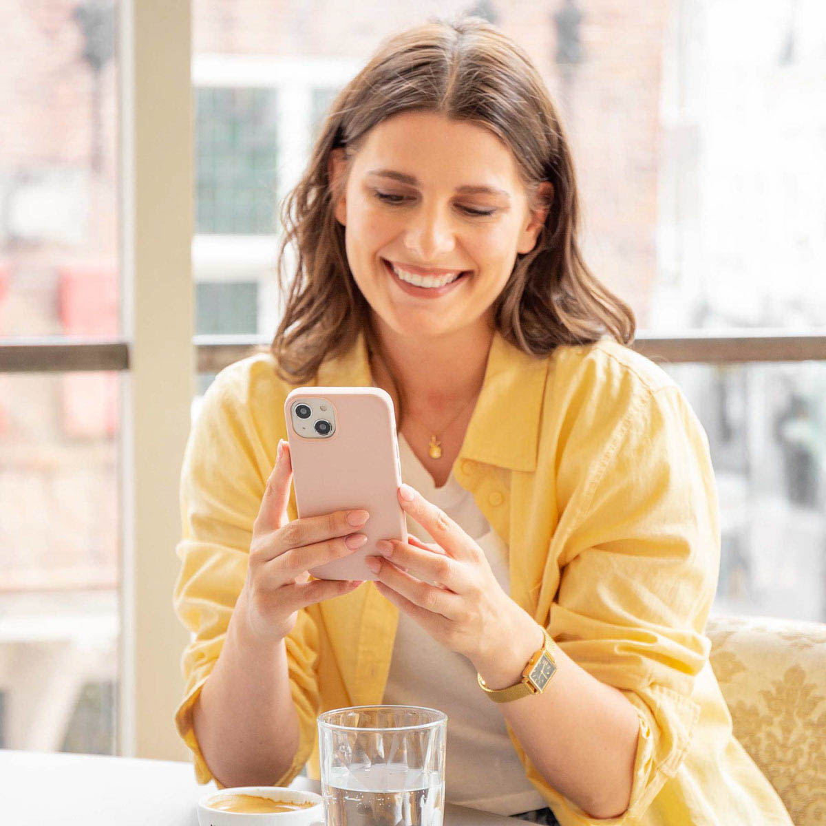 Uśmiechnięta kobieta w żółtej koszuli siedzi w środku i patrzy na różowy smartfon.
