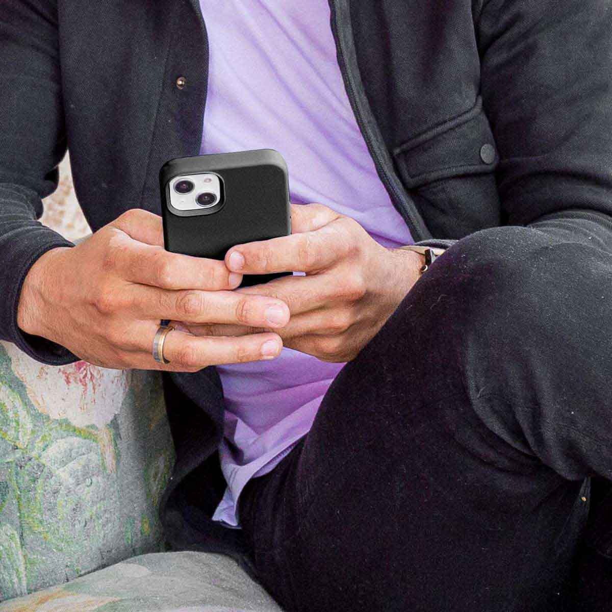 Osoba w czarnej kurtce i fioletowym t-shircie siedzi i trzyma w obu rękach czarny smartfon.