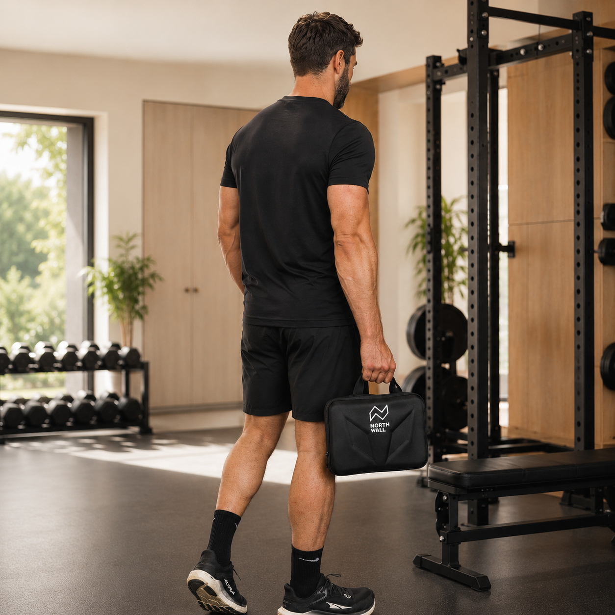 Ein Mann im schwarzen Sportoutfit steht im Fitnessstudio und hält einen kleinen Koffer.
