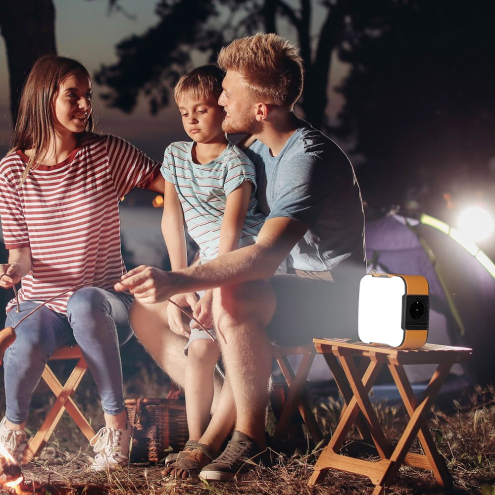 Una familia se sienta junto a una fogata al atardecer, con una luz de camping portátil sobre un taburete.