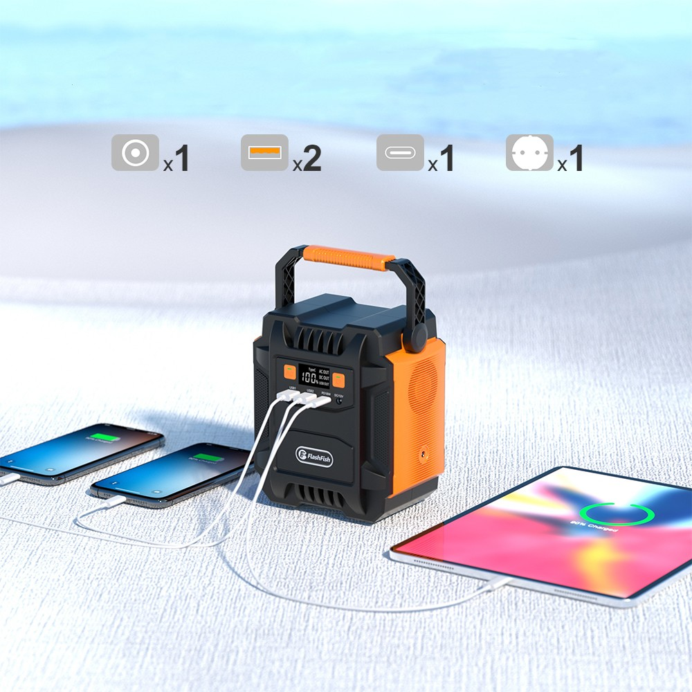 Una estación de energía portátil negra y naranja cargando dos smartphones y una tablet en una playa de arena.