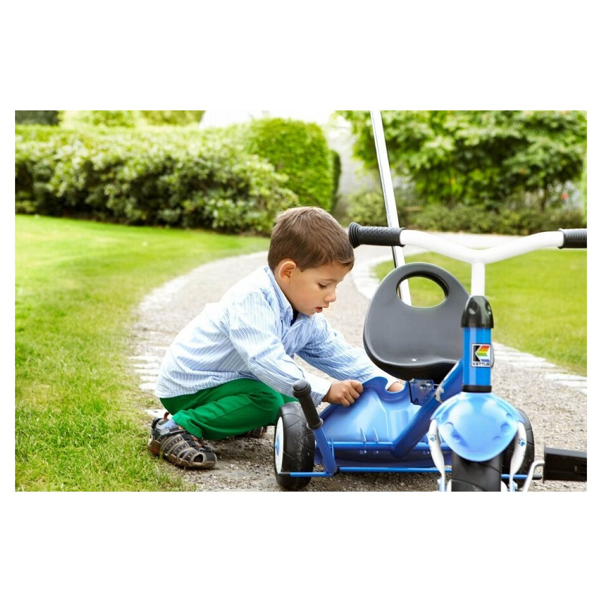 Kleiner Junge kniet auf einem Gartenweg und repariert ein blaues Dreirad.