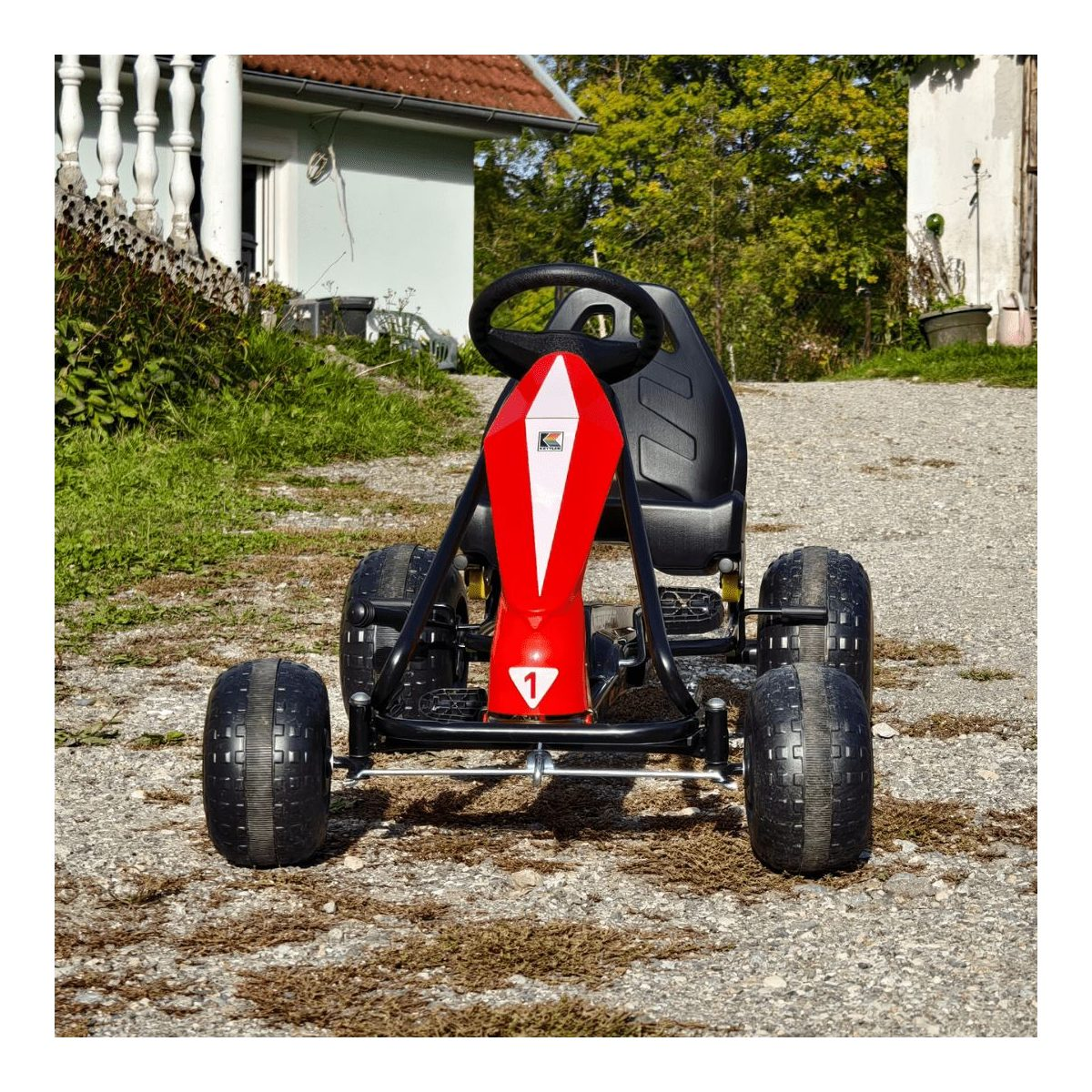 Ein rotes und schwarzes Kinder-Go-Kart steht auf einem Schotterweg vor einem Haus an einem sonnigen Tag.