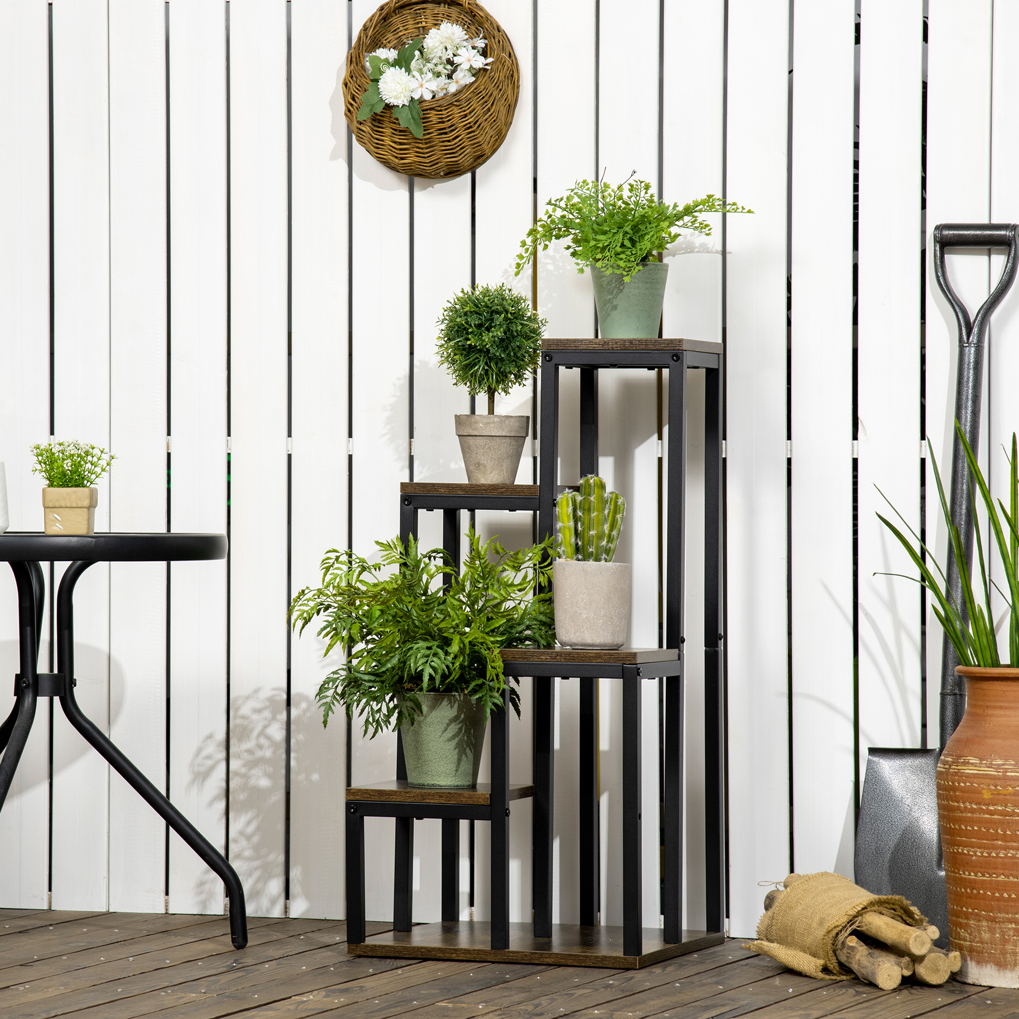 Un soporte para plantas de varios niveles con varias macetas contra una cerca de madera blanca, con pala y cesta.
