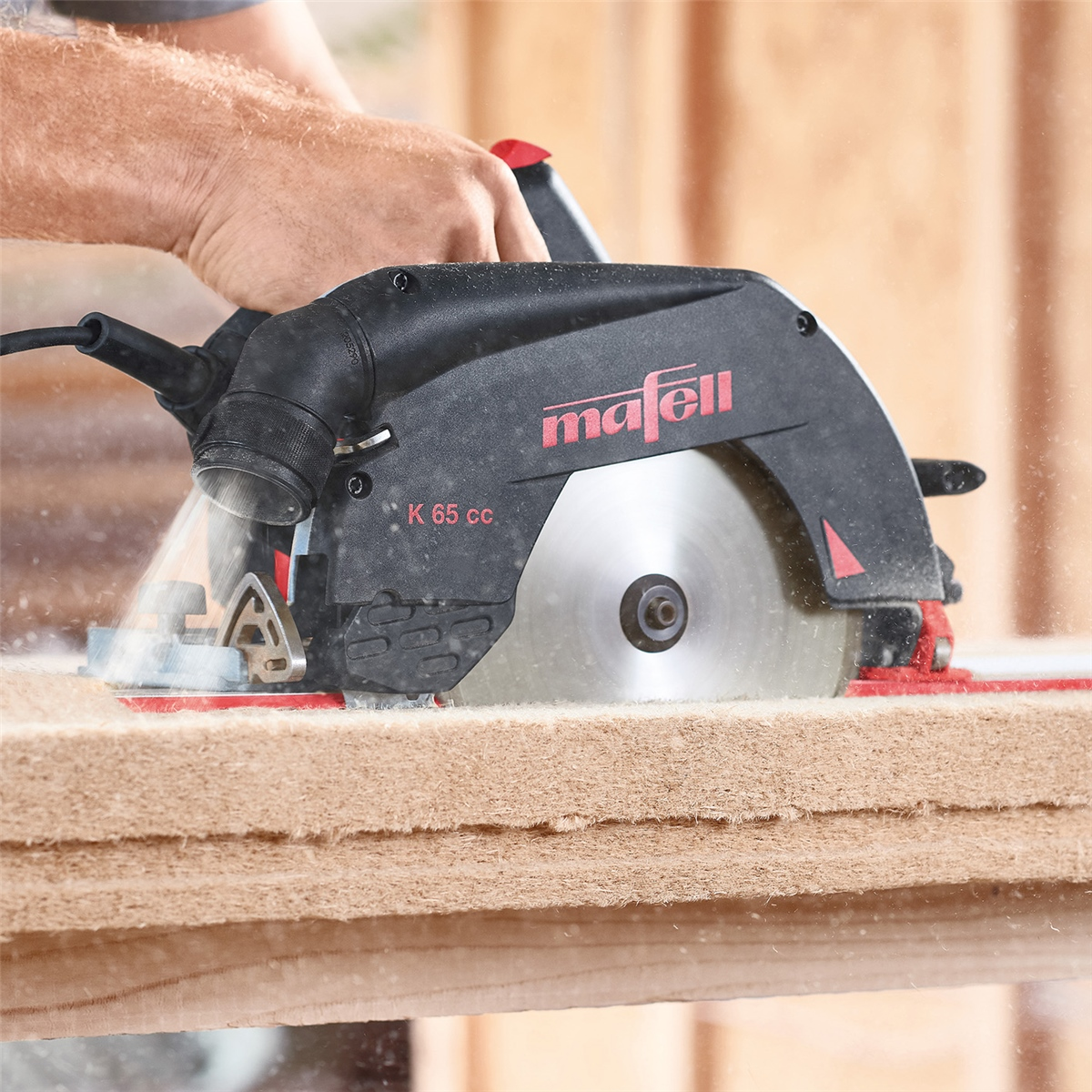 Eine Hand bedient eine schwarze Mafell K 65 cc Handkreissäge, die ein dickes Holzbrett schneidet.
