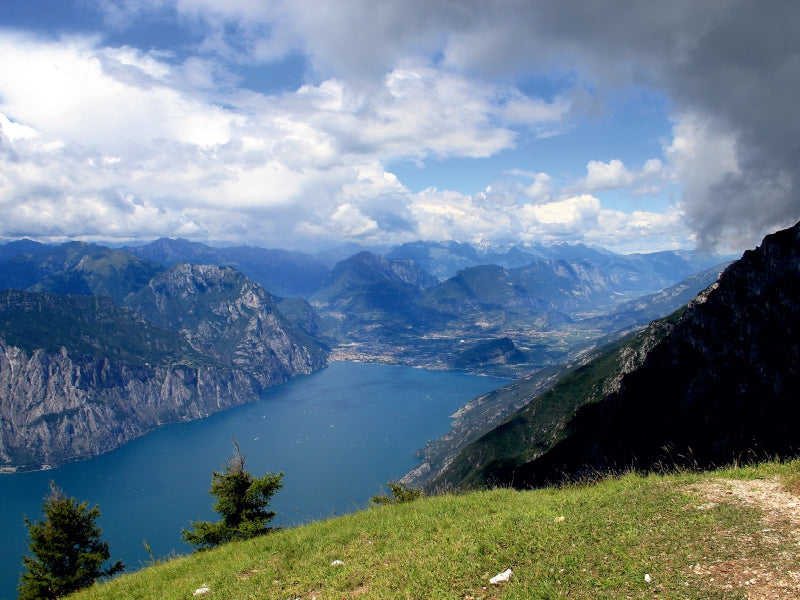 Blick von einem Berg auf einen blauen See, umgeben von steilen Felsen, Tälern und einem bewölkten Himmel.