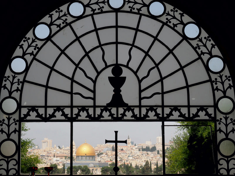 Blick durch ein verziertes Bogenfenster mit Kreuz auf die goldene Kuppel des Felsendoms in Jerusalem und die Stadt.
