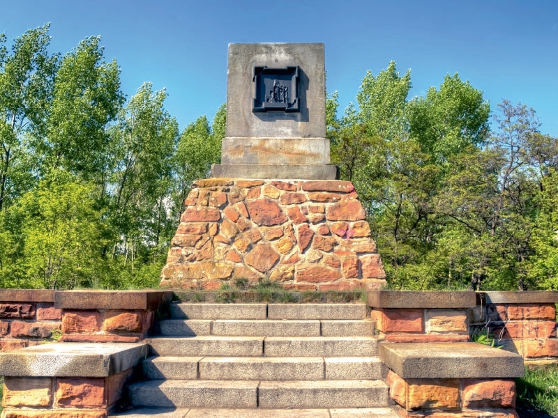 Ein Steindenkmal mit bronzenem Relief auf einem Steinsockel, umgeben von grünen Bäumen unter blauem Himmel.