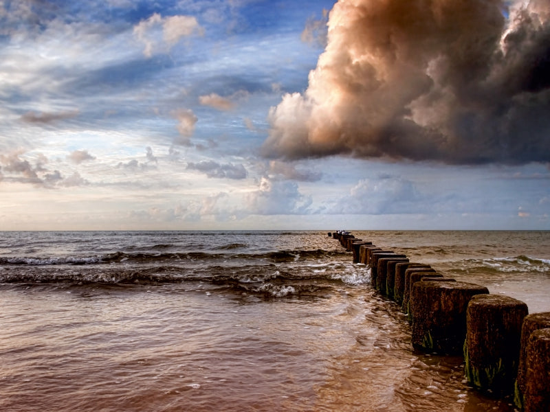 Hölzerne Buhnen ragen bei Sonnenuntergang unter einem dramatischen Himmel mit dunklen Wolken ins ruhige Meer.