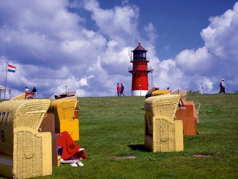 Gelbe Strandkörbe auf einer grünen Wiese mit einem roten Leuchtturm im Hintergrund unter blauem Himmel.
