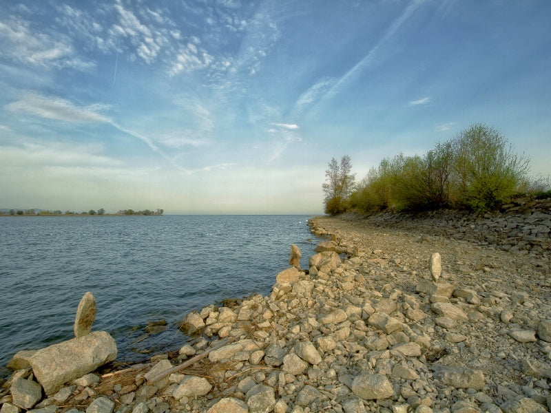 Felsiges Seeufer mit ruhigem Wasser unter einem blauen, wolkenverhangenen Himmel.