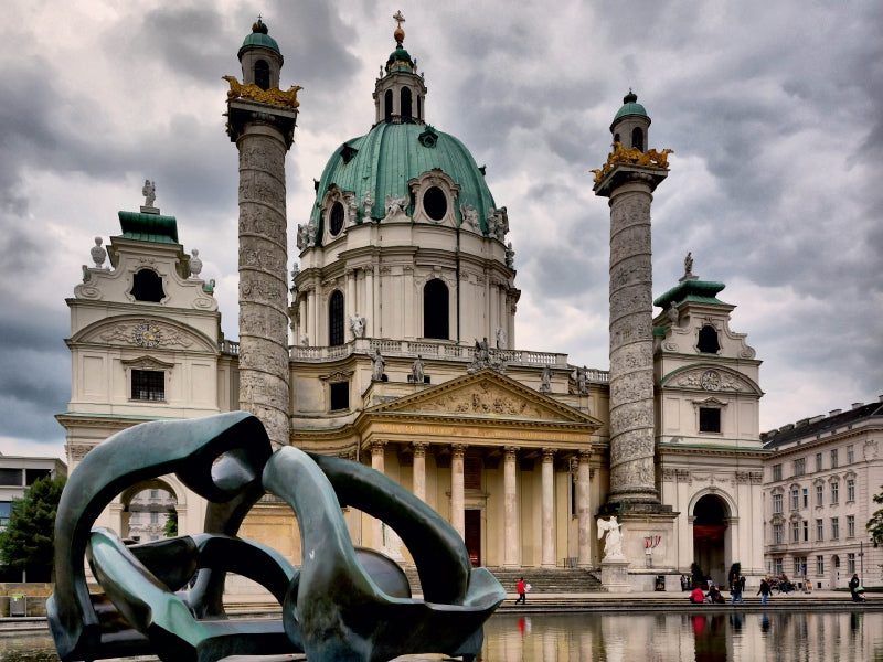 Barocke Karlskirche in Wien mit grüner Kuppel, zwei großen Säulen und einer abstrakten Skulptur vorne am Wasser.