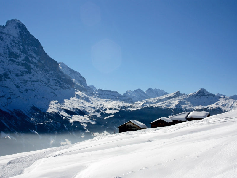 Verschneite Holzhütten vor einer alpinen Bergkette mit blauem Himmel.