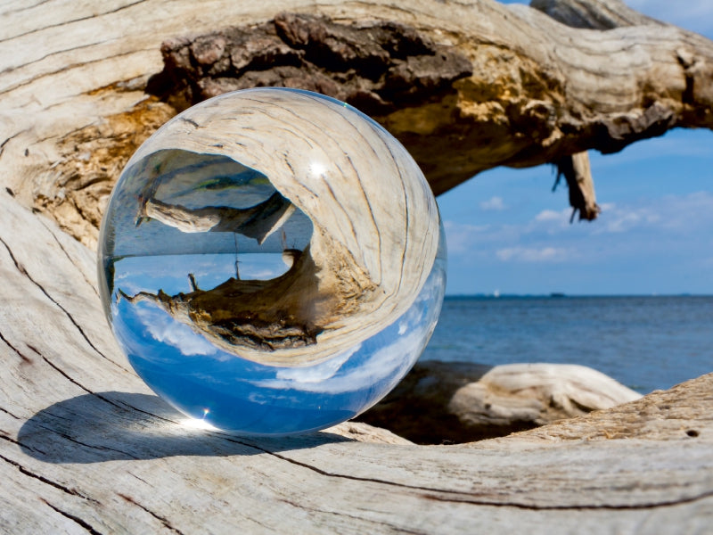 Eine glasklare Kugel auf Treibholz spiegelt die Umgebung, den blauen Himmel und das Meer verkehrt herum.