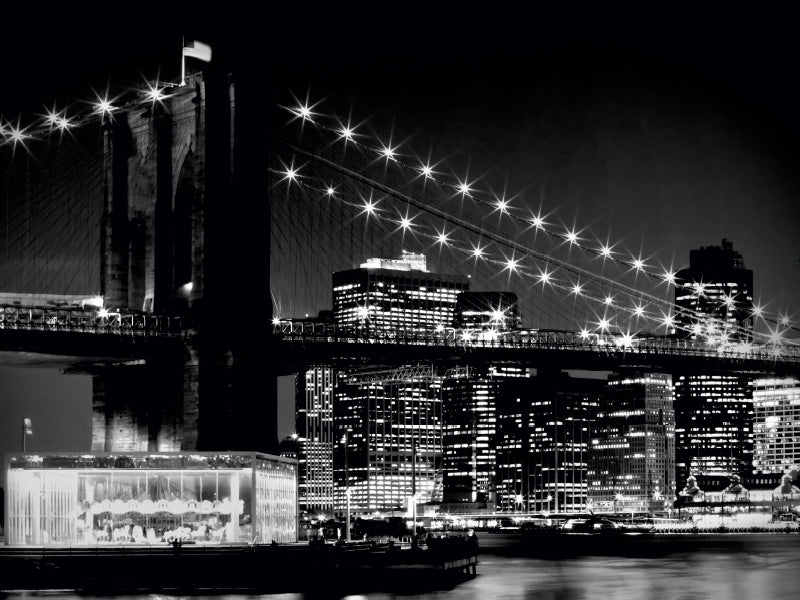 Schwarz-Weiß-Ansicht der Brooklyn Bridge nachts mit glitzernden Lichtern, beleuchteten Wolkenkratzern und einem hellen Karussell im Vordergrund.
