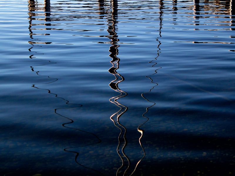 Wellenförmige Reflexionen von Holzpfählen auf der dunklen, blauen Wasseroberfläche eines Sees.