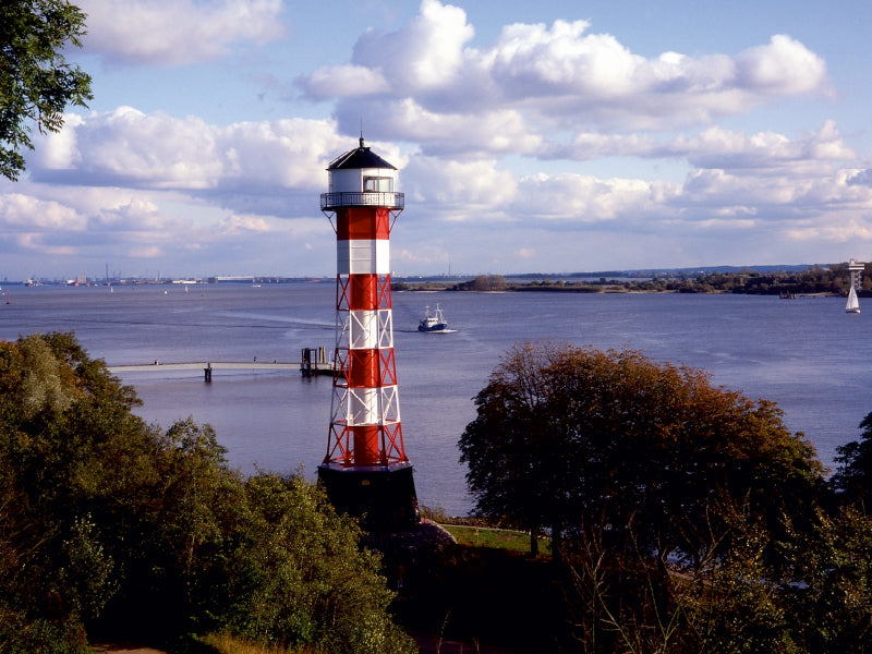 Rot-weißer Leuchtturm am Wasser mit Bäumen im Vordergrund und Booten auf dem Fluss unter bewölktem Himmel.