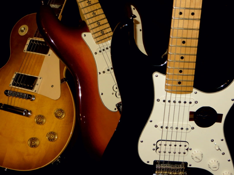 Drei E-Gitarren mit unterschiedlichen Oberflächen und Pickguards stehen vor einem schwarzen Hintergrund.