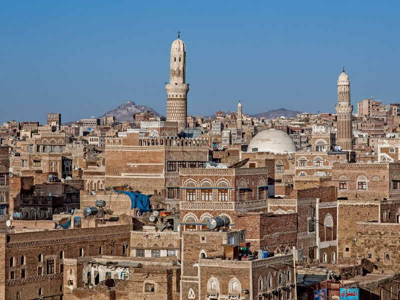 Blick über die alte Stadt Sanaa mit traditionellen Lehmziegelgebäuden, Minaretten und Bergen im Hintergrund.