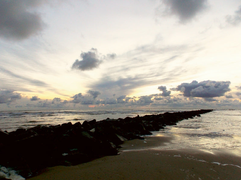 Eine steinige Buhne ragt bei Sonnenuntergang unter bewölktem Himmel ins Meer.