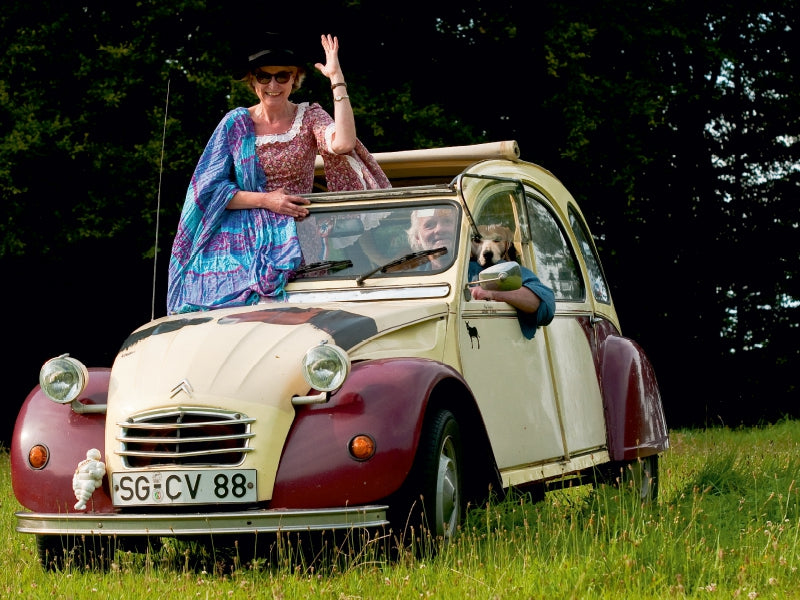 Eine Frau in historischer Kleidung und ein Mann mit Hund sitzen in einem bunt bemalten Oldtimer auf einer Wiese.