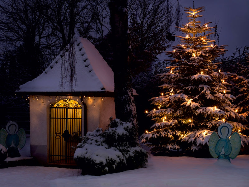 Eine schneebedeckte Kapelle und ein festlich beleuchteter Weihnachtsbaum im Schnee bei Nacht.