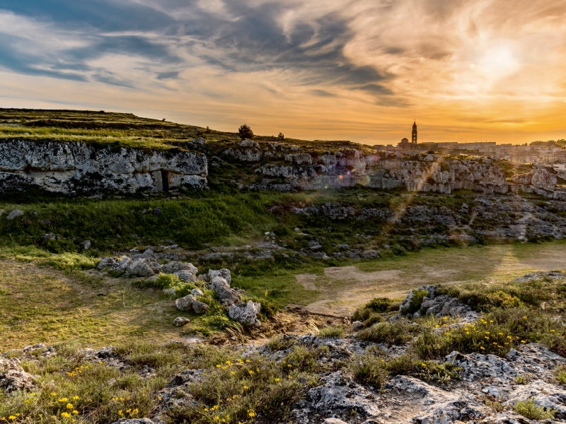Landschaft mit felsigen Klippen und gelben Blumen im Vordergrund unter einem dramatischen Sonnenuntergangshimmel.