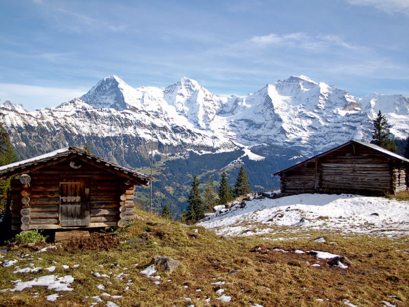 Zwei Holzhütten auf einer verschneiten Wiese vor einer majestätischen schneebedeckten Bergkette.