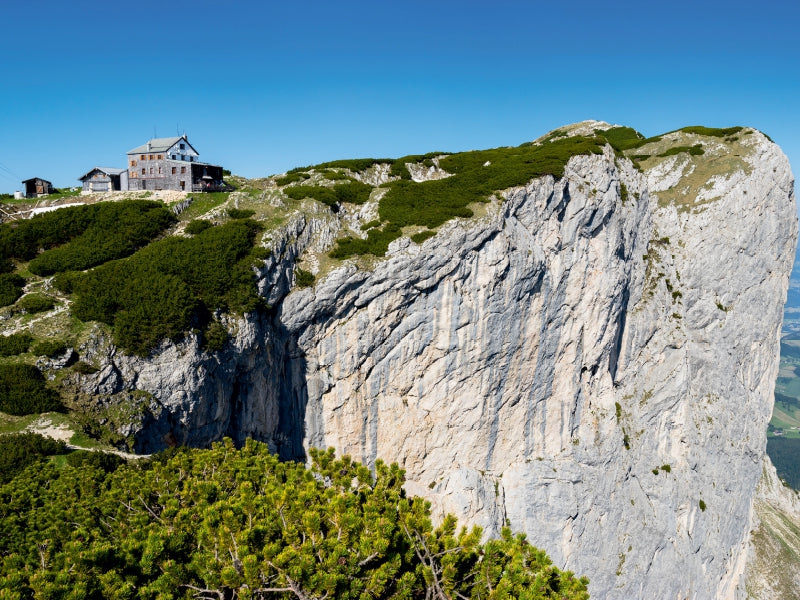 Eine Berghütte steht auf einem steilen Felsen unter blauem Himmel, umgeben von grünen Büschen.