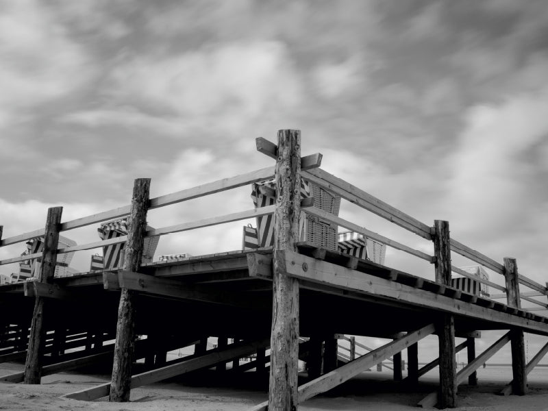 Schwarzweißfoto eines hölzernen Strandpavillons auf Stelzen mit gestreiften Strandkörben gegen bewölkten Himmel.