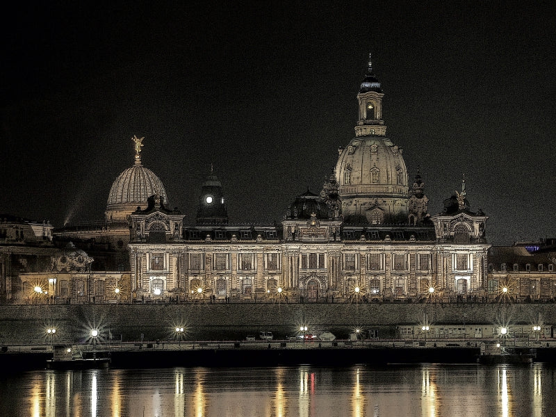 Nächtliche Ansicht der Dresdner Frauenkirche und des Lipsius-Baus am Flussufer mit hellen Lichtern.