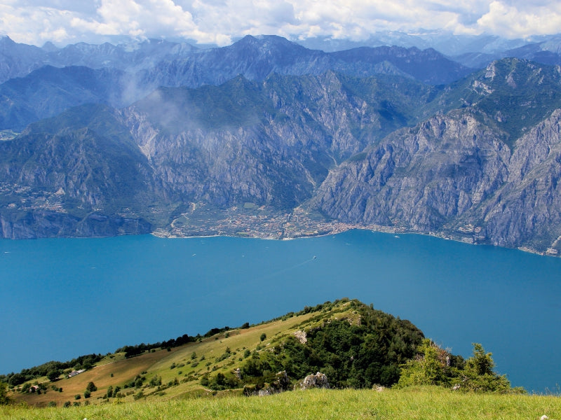 Blick von oben auf einen tiefblauen See, umgeben von steilen Bergen und einem grünen Voralpenhang im Vordergrund.