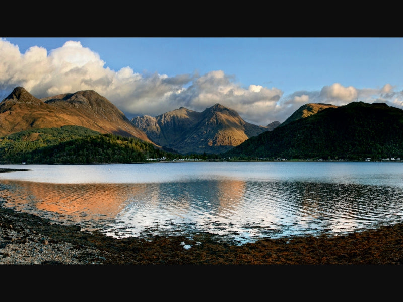 Ruhiger See mit bewaldeten Ufern und hoch aufragenden Bergen unter einem wolkenverhangenen blauen Himmel.
