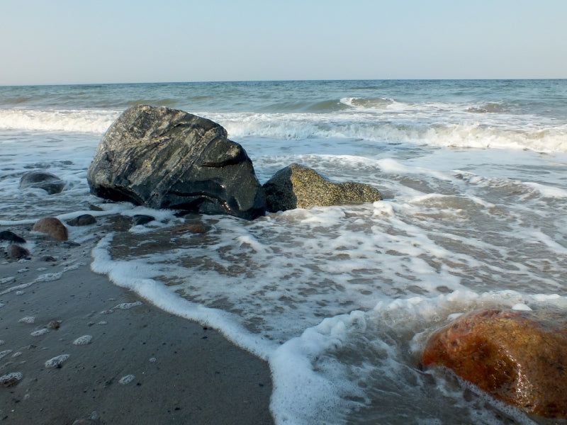 Wellen brechen an einem Sandstrand mit dunklen und rötlichen Felsen im Vordergrund unter klarem Himmel.