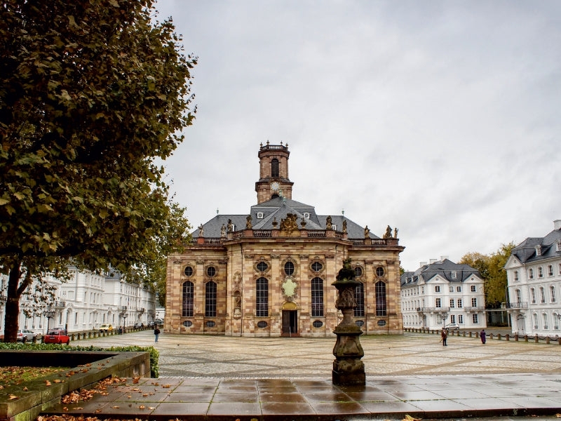 Große historische Barockkirche auf einem Platz, umgeben von weißen Gebäuden und Bäumen.