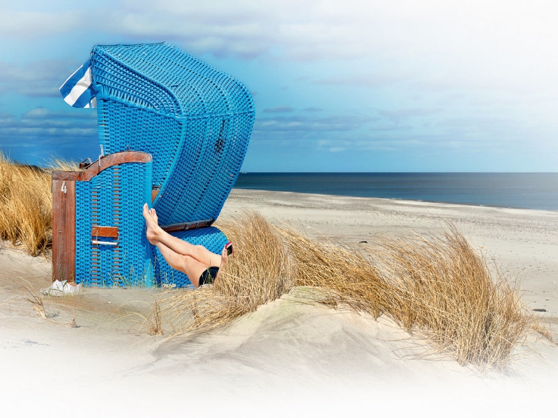 Eine Person entspannt in einem blauen Strandkorb in den Dünen am Meer unter bewölktem Himmel.