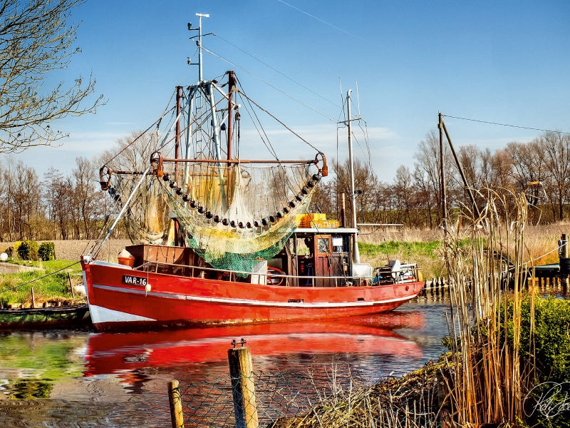 Ein rotes Fischerboot mit Netzen liegt auf einem ruhigen Kanal, umgeben von Schilf und Bäumen unter blauem Himmel.