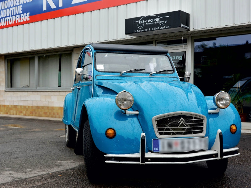Ein hellblaues klassisches Auto steht vor einer Werkstatt mit Schildern und Fenstern.