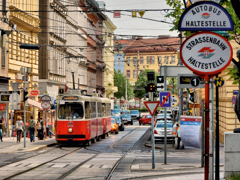 Eine rote Straßenbahn fährt auf einer städtischen Straße mit Schildern im Vordergrund und Gebäuden im Hintergrund.