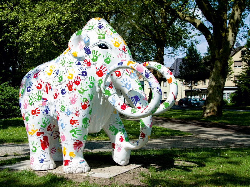 Eine weiße Mammut-Statue, bedeckt mit bunten Handabdrücken und Namen, steht in einem sonnigen Park.