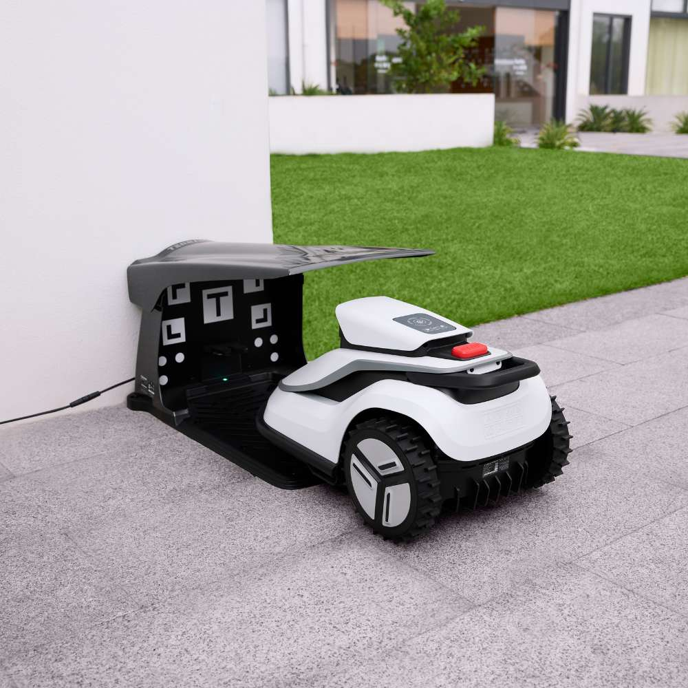Un robot tondeuse blanc et noir est garé sur une terrasse devant sa station de charge noire près d'un mur blanc.