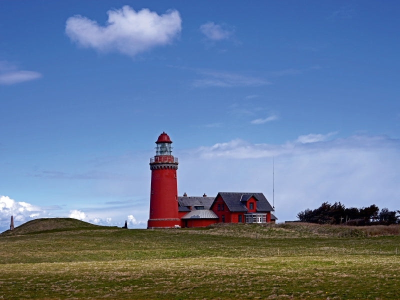 Ein roter Leuchtturm und angrenzendes Gebäude auf grüne Wiese unter blauem Himmel mit Wolken.