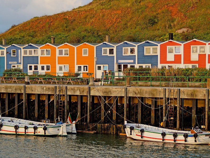 Bunt getäfelte Häuser stehen an einem Pier mit vertäuten Booten und einer grünen Klippe im Hintergrund.