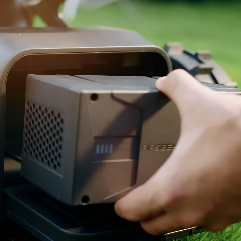 Une main insère une batterie grise dans un appareil, avec de l'herbe verte en arrière-plan.
