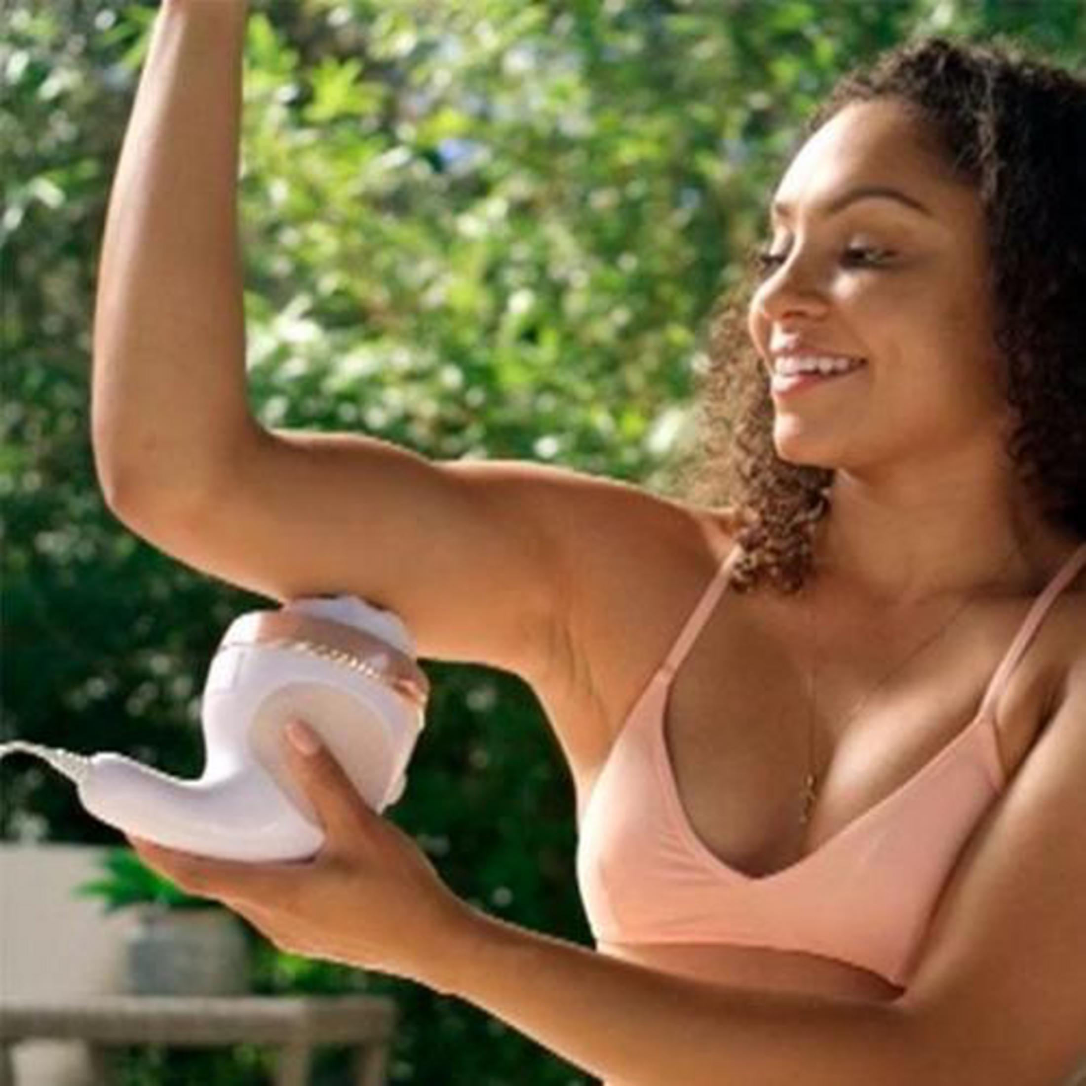Une femme souriante en haut pêche tient un appareil blanc et or rose près de son aisselle à l'extérieur.
