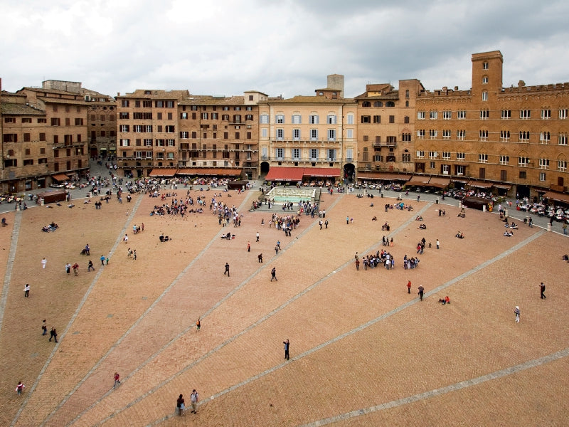 Ein großer, teilgepflasterter Marktplatz in einer italienischen Stadt, umgeben von alten roten Backsteingebäuden.