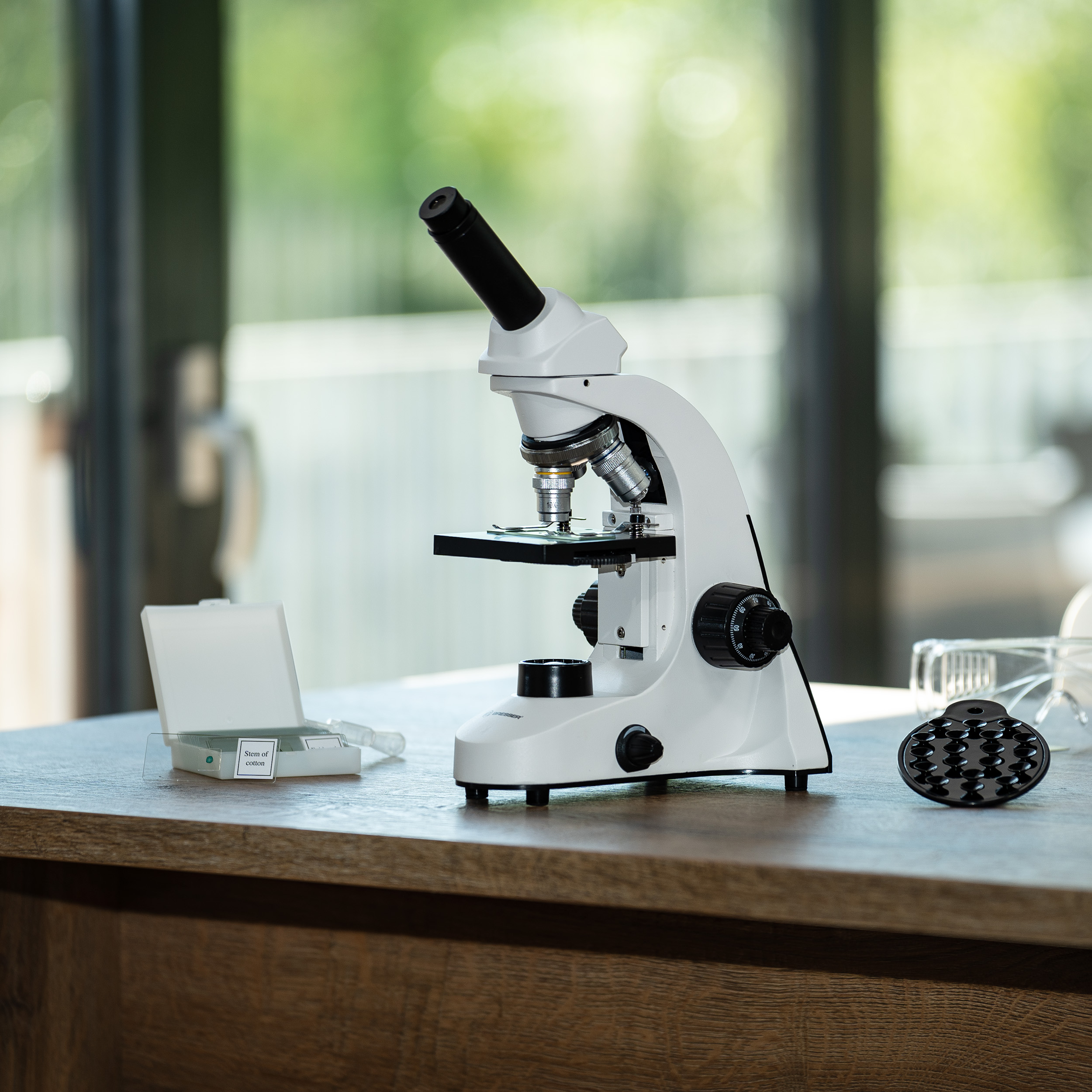 Un microscopio blanco en un escritorio de madera con una caja de portaobjetos y accesorios, con fondo borroso.