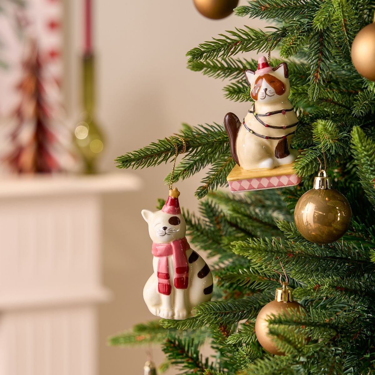 Deux ornements de chats en céramique avec bonnets et écharpes pendent sur un sapin avec des boules dorées.
